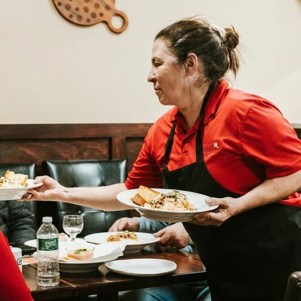 A server in a red shirt and black apron delivers plates of food to customers at a restaurant table.