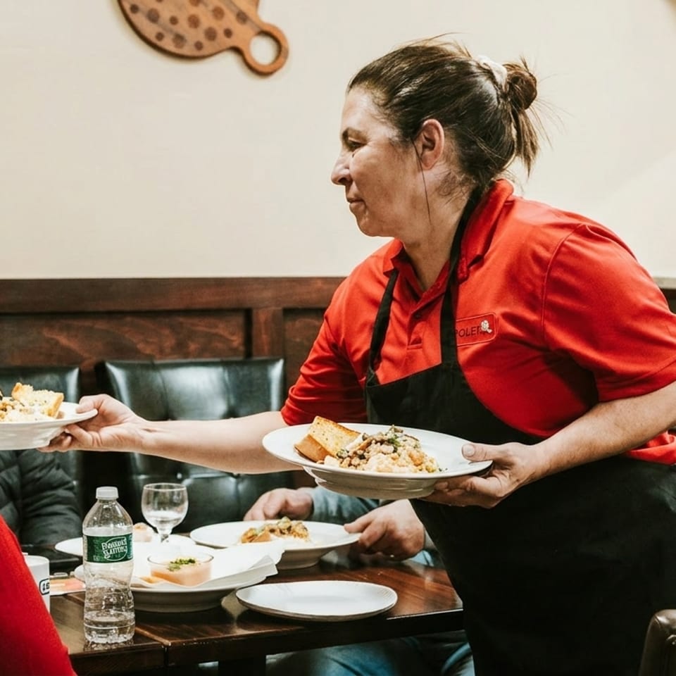 A server in a red shirt and black apron delivers plates of food to customers at a restaurant table.