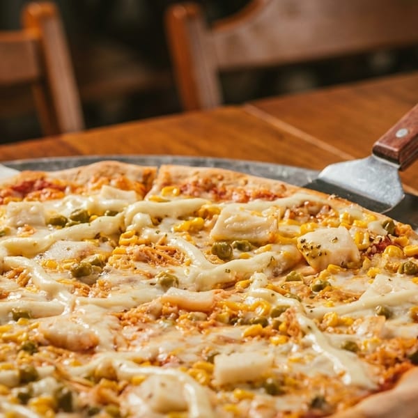 A freshly baked pizza with corn, peas, and cheese on a metal tray with a server.